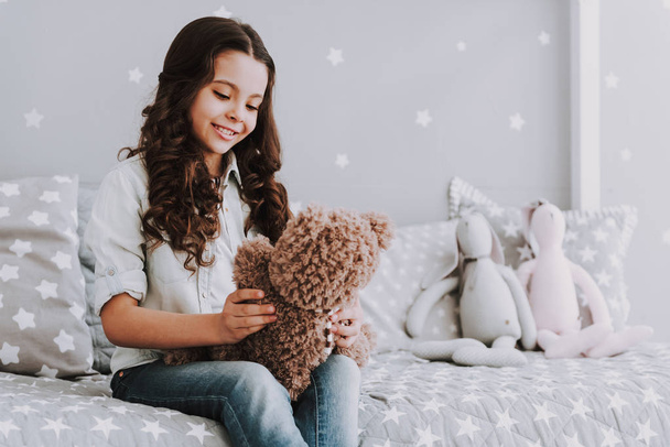 Cute Little Girl Sitting on Bed holding Teddy Bear. Smiling Adorable Little Girl Sitting on Bed and Holding Bear Toy at Home. Beautiful Little Girl in Bedroom. Childhood Concept - Foto, afbeelding