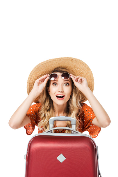 excited beautiful girl holding sunglasses and sitting near travel bag isolated on white  - Photo, Image