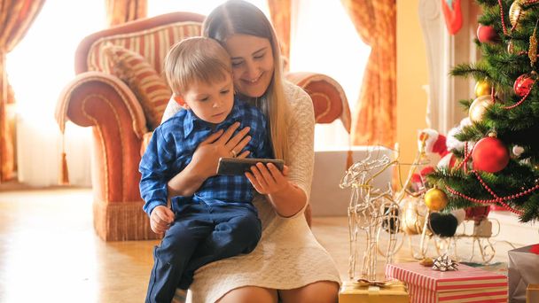 Totyogó fiú wathcing video-ra mozgatható telefon-val anya: nappali portréja díszített karácsonyra - Fotó, kép