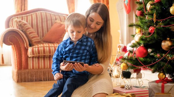 Portret van de moeder met peuter jongen ontspannen op kerstochtend en kijken naar video op mobiele telefoon - Foto, afbeelding