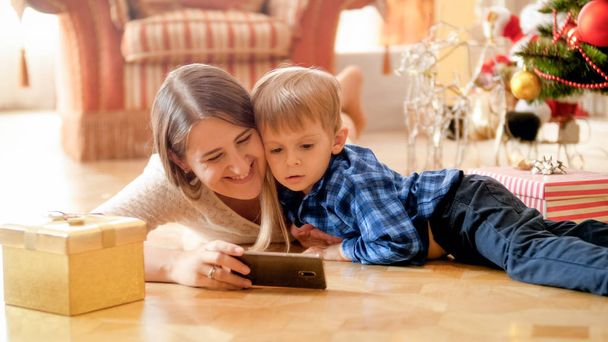 Portret matki szczęśliwy uśmiechający się z toddler chłopiec leżącego w salonie pod choinkę i oglądanie filmów na smartphone - Zdjęcie, obraz