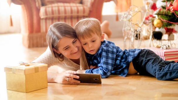 Maluch mały chłopiec z matka oglądanie bajki na telefon na Boże Narodzenie rano - Zdjęcie, obraz