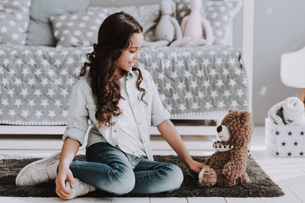 Cute Little Girl Sitting on Carpet with Teddy Bear. Smiling Adorable Little Girl Sitting on Crpet next to Bed and Playing with Bear Toy at Home. Beautiful Little Girl in Bedroom. Childhood Concept - 写真・画像