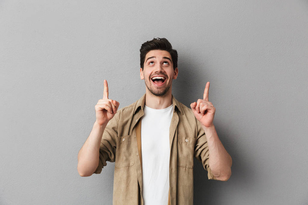 Portrait of a cheerful young casual man pointing up at copy space isolated over gray background - Foto, Imagen