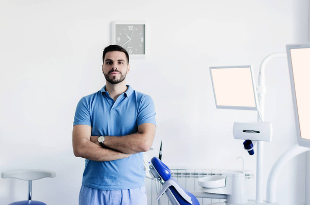 Πορτρέτο του ένα ευτυχισμένο νεαρό επαγγελματία οδοντίατρο αρσενικό, χαμογελά στη φωτογραφική μηχανή που παρουσιάζουν περήφανα στο οδοντικό γραφείο του. - Φωτογραφία, εικόνα