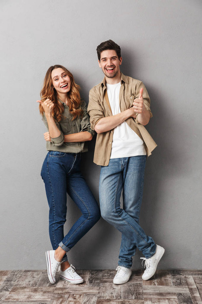 Full length portrait of a cheerful young couple standing together showing thumbs up isolated over gray - Photo, image
