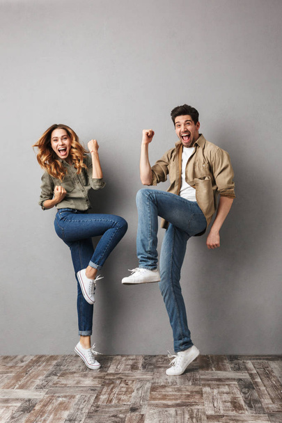 Full length portrait of a joyful young couple jumping together and celebrating success isolated over gray - 写真・画像