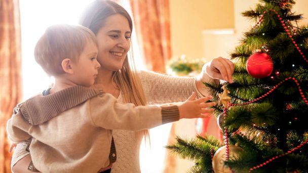 glücklich lächelnde Mutter mit kleinem Sohn beim Dekorieren des Weihnachtsbaums mit Kugeln - Foto, Bild