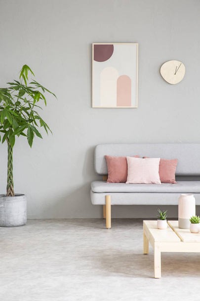 Poster and clock above settee with pink pillows in grey flat interior with plant and table. Real photo - Foto, afbeelding
