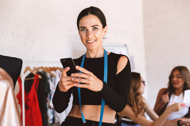 Εύθυμη νεαρή γυναίκα ρούχα σχεδιαστών στο atelier studio λειτουργεί με συναδέλφους σε φόντο, κρατώντας το κινητό τηλέφωνο - Φωτογραφία, εικόνα