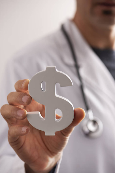 Nahaufnahme eines jungen kaukasischen Arztes mit einem Dollarsymbol in der Hand, das die Konzepte der Gesundheitsindustrie oder die Kosten des Gesundheitswesens darstellt - Foto, Bild
