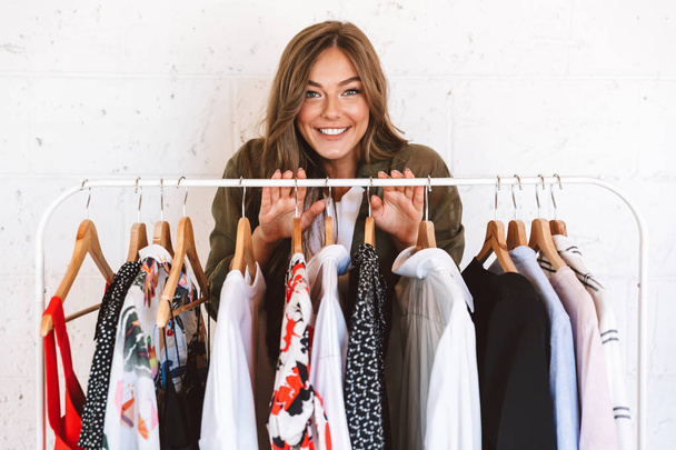 Lovely young woman clothes designer standing at the clothes rack indoors - Photo, image