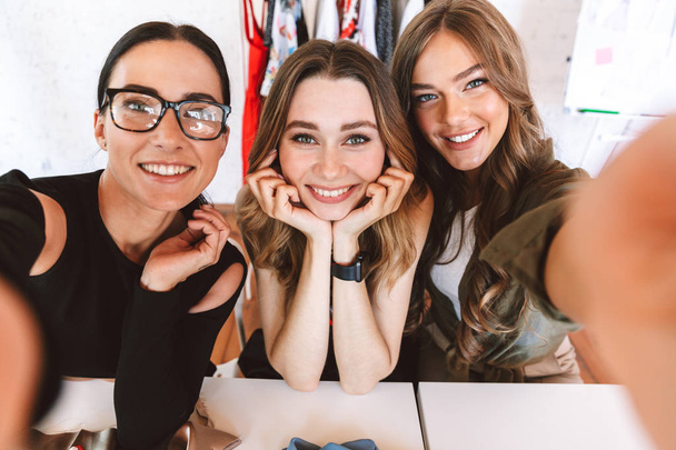 Three attractive young women clothes designers working together at the atelier, sitting at the table and taking selfie - Photo, Image