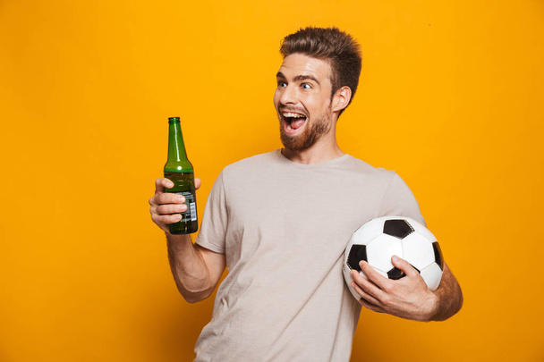 Image of handsome man standing isolated over yellow wall backgroung drinking beer holding football. - Foto, Bild