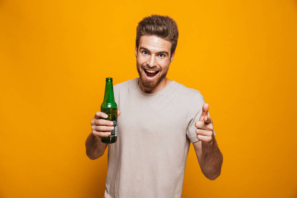 Image of handsome man standing isolated over yellow wall backgroung drinking beer pointing to you looking at camera. - Photo, Image