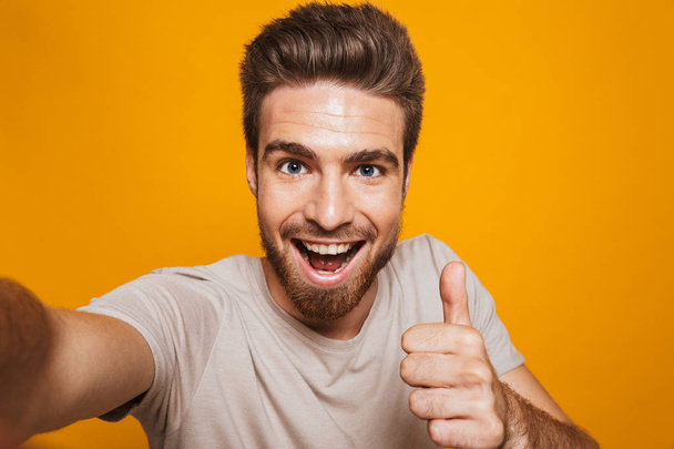 Image of handsome happy man standing isolated over yellow wall backgroung make a selfie by camera with thumbs up. - Valokuva, kuva