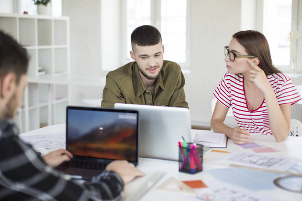 Молодой человек в рубашке и женщина в полосатой футболке и очках работают вместе с ноутбуком. Группа классных парней, проводящих время в современном офисе
  - Фото, изображение