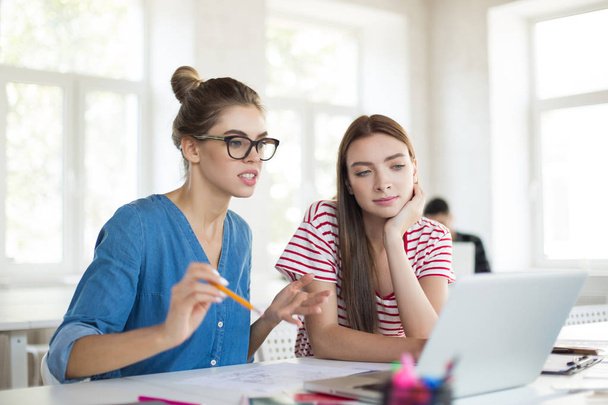 Όμορφο κορίτσι σε γυαλιά με μολύβι σκεπτικά συνεργάζονται με συνάδελφο για φορητό υπολογιστή. Νεαρές γυναίκες αφιερώνουν χρόνο στο σύγχρονο γραφείο  - Φωτογραφία, εικόνα