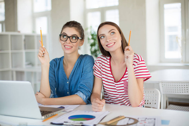 Lächelndes Mädchen in Jeanshemd und Brille und Mädchen im gestreiften T-Shirt mit Bleistiften in den Händen, die fröhlich nach oben schauen. junge Frauen arbeiten mit Laptop im modernen Büro zusammen - Foto, Bild