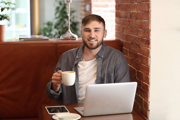 Young freelancer drinking coffee while working on laptop in cafe - Фото, изображение