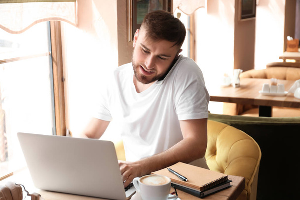 Fiatal szabadúszó beszél mobiltelefonon, miközben dolgozik a laptop a kávézóban - Fotó, kép