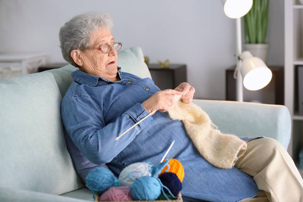 Seniorin sitzt zu Hause auf Sofa und strickt Pullover - Foto, Bild