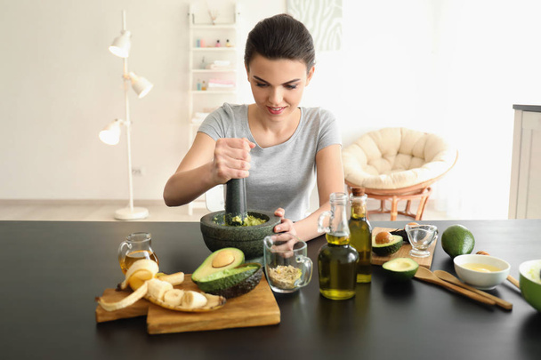 Young woman making nourishing mask with avocado in kitchen - Photo, image