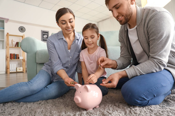 Happy mała dziewczynka z rodzicami, siedząc na dywanie i wprowadzenie monet w pomieszczeniu Skarbonka. Koncepcja oszczędności pieniądze - Zdjęcie, obraz