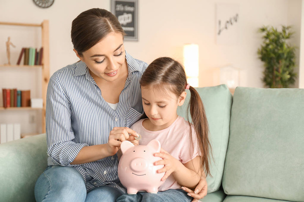 Little girl with her mother sitting on sofa and putting coin into piggy bank. Money savings concept - Фото, зображення