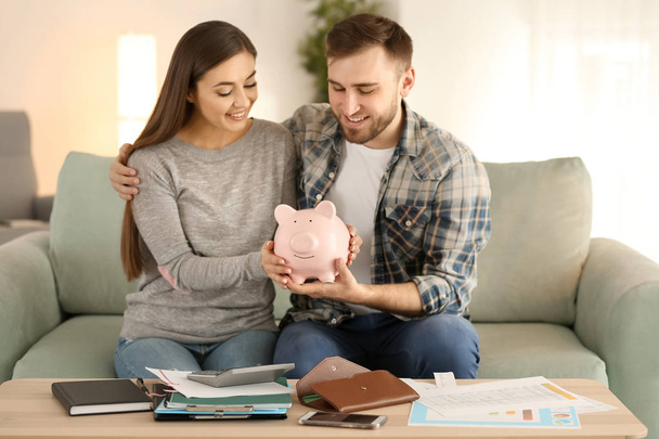Happy couple sitting on sofa and holding piggy bank indoors. Money savings concept - 写真・画像