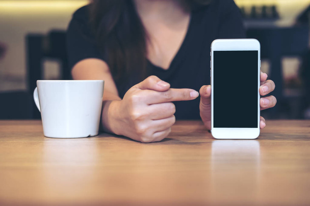 Attrappe einer schönen Frau, die ein weißes Handy mit leerem schwarzen Bildschirm und weißer Kaffeetasse auf einem Holztisch im Restaurant hält, zeigt und zeigt  - Foto, Bild