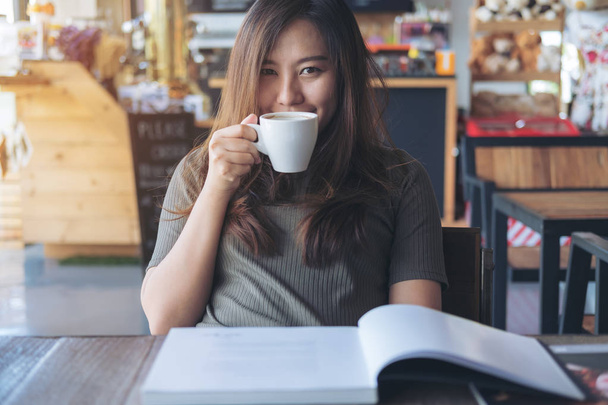 Gros plan image d'une belle femme asiatique lisant un livre tout en tenant et en buvant du café chaud dans un café moderne
  - Photo, image