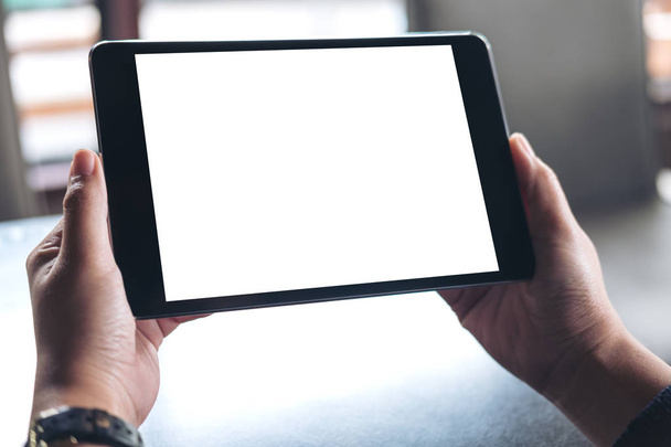 Макетне зображення рук, що тримають чорний планшетний ПК з білим порожнім екраном на дерев'яному фоні столу в кафе
 - Фото, зображення