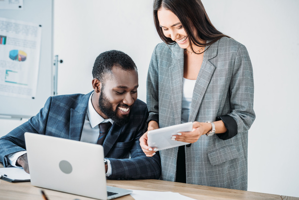 smiling african american businessman and asian businesswoman looking at tablet in office  - Foto, Bild
