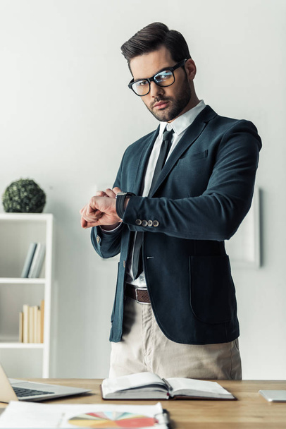 Bonito homem de negócios em óculos está olhando para seu relógio enquanto está em seu local de trabalho no escritório
 - Foto, Imagem