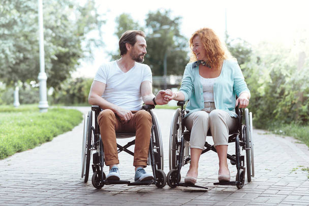 Loving mature couple holding hands together while sitting in wheelchairs - Фото, зображення