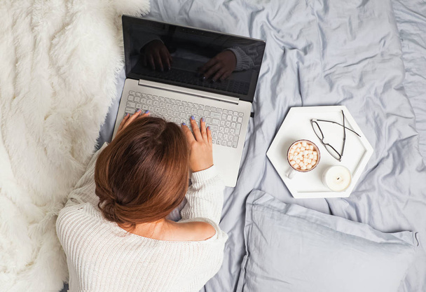 Молодая женщина в белом свитере лежит на кровати и использует ноутбук
. - Фото, изображение