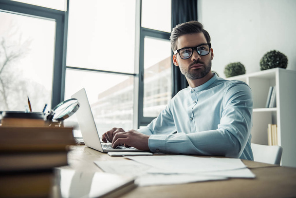 Handsome pensive businessman in eyeglasses is looking away and thinking while working with the laptop in the office - Foto, Imagen