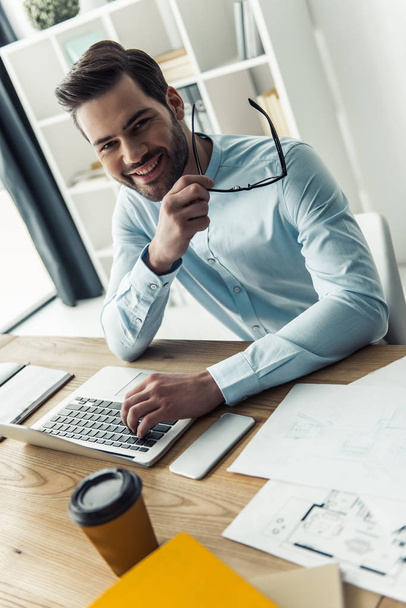 Hombre de negocios guapo está sosteniendo gafas, mirando a la cámara y sonriendo mientras trabaja con el ordenador portátil en la oficina
 - Foto, imagen