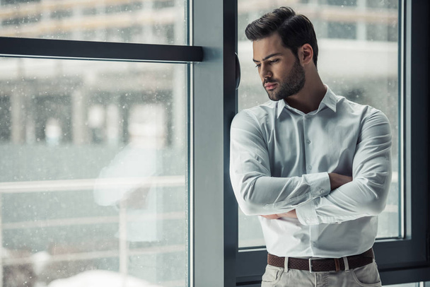 Hombre de negocios pensativo guapo está mirando por la ventana, frotando su barbilla y pensando mientras está de pie en la oficina
 - Foto, imagen