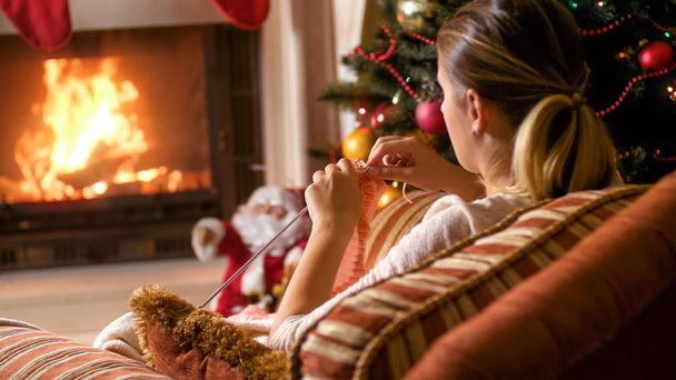 Young woman knitting woolen scarf next to fireplace and Christmas tree - Fotografie, Obrázek
