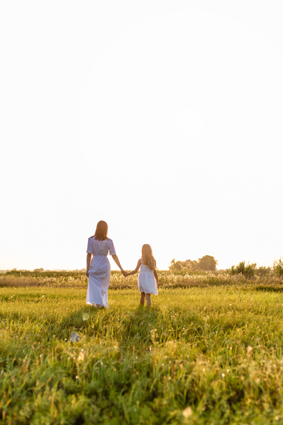 rear view of mother and daughter holding hands and walking by green meadow with sunset sky on background - Photo, Image