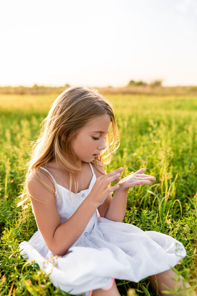 adorable little child sitting on green grass and looking at hand under sunset rays - Foto, Imagem