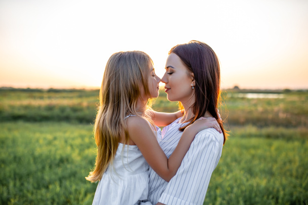close-up portrait of beautiful mother and daughter embracing and touching noses in green field on sunset - Foto, imagen