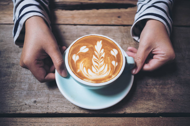 Immagine da primo piano delle mani della donna che tengono e mostrano una tazza blu di caffè latte caldo con arte del latte su tavolo vintage in legno - Foto, immagini