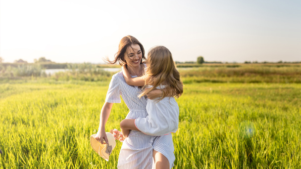 beautiful happy mother and daughter embracing in green field on sunset - Fotografie, Obrázek