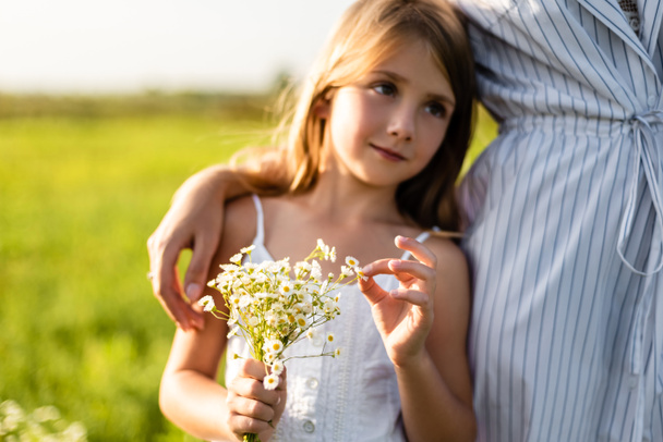 cropped shot of mother embracing daughter while she holding field flowers bouquet in green meadow - Фото, зображення