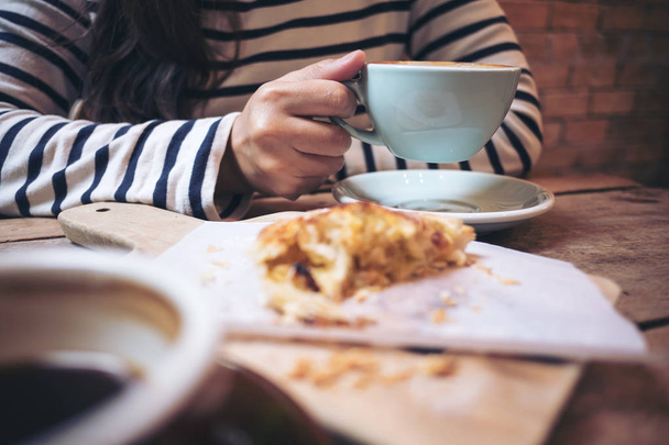 Closeup image of a woman holding and drinking hot latte coffee with a piece of raisin danish on wooden vintage table in coffee shop - Photo, Image