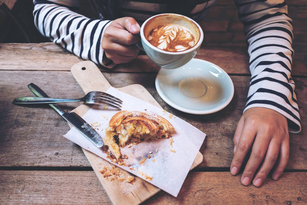Top view image of a woman holding and drinking hot latte coffee with a piece of raisin danish on wooden vintage table in coffee shop - Photo, Image