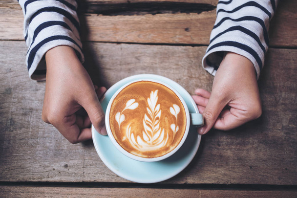 Top view image of woman's hands holding and showing a blue cup of hot latte coffee with latte art on wooden vintage table - Photo, Image